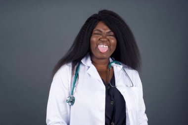 Vücut dili. Gri duvara karşı poz veren, suratını asan, bir şeye karşı hoşnutsuzluk gösteren güzel bir Afrikalı Amerikalı kadın doktor..