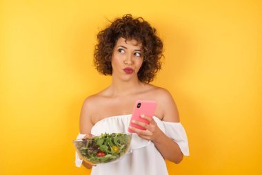 Düşünen genç ve güzel bir kadının salata tuttuğunu hayal et. Sıradan giyinmiş, parlak duvar arkasında izole bir şekilde cep telefonu kullanarak poz veriyor..