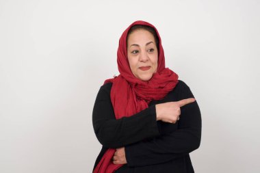 Olgun esmer bir kadın kamerada poz veriyor, hileli bir bakış ve yana hareket ediyor. Bayan tasarımcı, işaret parmağıyla harika bir ürün sunuyor. Jest konsepti. Stüdyoda poz veren başörtüsü takan orta yaşlı bir Arap kadın.