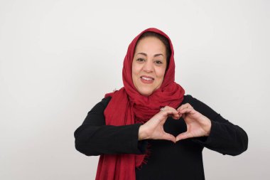 Tecrit edilmiş bir geçmişi olan olgun bir kadın. Elleriyle kalp sembolü ve şekli gösteren bir aşkla gülümsüyor. Romantik bir kavram. Stüdyoda poz veren başörtüsü takan orta yaşlı bir Arap kadın.