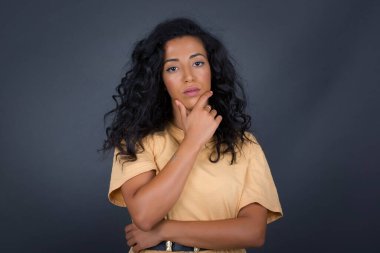 Düşünceli gülümseyen kızın portresi elini çenesinin altında tutuyor, doğrudan kameraya bakıyor, ilgiyle karışık bir şey dinliyor, sıradan giyiniyor, gri duvara karşı poz veriyor. Gençlik kavramı.