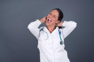 Güven ve cinsel ilişki konsepti. Gri arka planda izole edilmiş, kendinden emin bir ifadeyle gülümseyen ve ellerini başının üstünde tutan çekici genç doktor portresi. Gri arkaplanda duruyor.