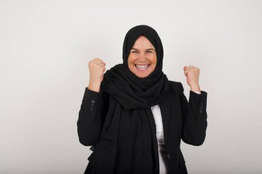 Başarılı, orta yaşlı, tesettürlü, tesettürlü, kollarını sıkan, sevinç ve heyecanla haykıran Müslüman bir kadının belden yukarı portresi. Zafer, başarı ve başarı kavramı.