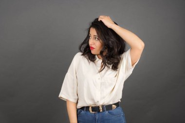 Güzel esmer kadın, sıradan giyinmiş, gözlerini kaçırıyor. Gri arkaplanda duruyor. 