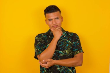 Düşünceli gülümseyen adamın portresi elini çenesinin altında tutuyor, doğrudan kameraya bakıyor, ilgi çekici bir şey dinliyor, sıradan giyiniyor, sarı duvara karşı poz veriyor..