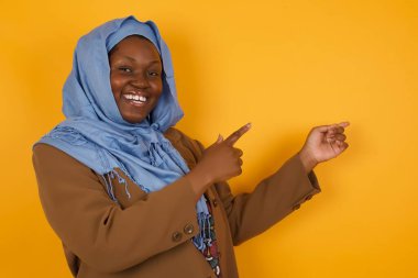 Reklam konsepti. Sarı arka planda gülümseyen Afro-Amerikan Müslüman kadın işaret parmağıyla işaret ediyor ve boş fotokopi alanına bakıyor..