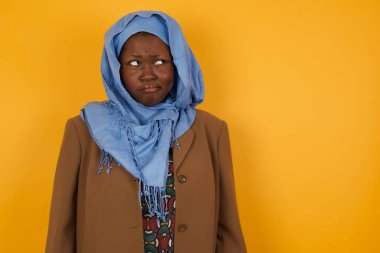 Şaşırmış genç Afrikalı Amerikalı Müslüman kadının fotoğrafı, kıvrımlı dudaklar ve endişeli bir bakış, önünde korkunç bir şey görüyor, sarı arka planda izole edilmiş.