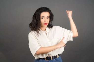 Güzel esmer kadın, sıradan giyinmiş, kaslarını gösteriyor. Gri arkaplanda duruyor. 