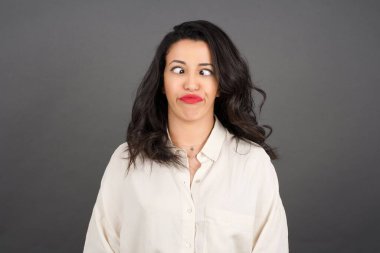 Güzel esmer kadın, kayıtsızca giyinmiş, gri arka planda komik bir ifade takınıyor..  