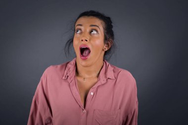 Şaşırmış esmer kadın omuz silkiyor, yana bakıyor, arkadaşlarıyla yeni planları için mutlu ve heyecanlı. Gri duvarda izole edilmiş..