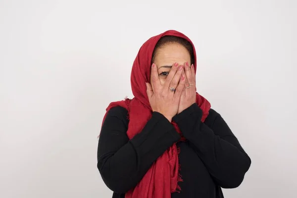 彼女の手で彼女の顔を覆う成熟した女性の概念的なイメージと屋内で立っている彼女の指の間の片目でピアス 何かや誰かから怖い スタジオでポーズするヘッドスカーフを身に着けている中年のアラブ女性 — ストック写真