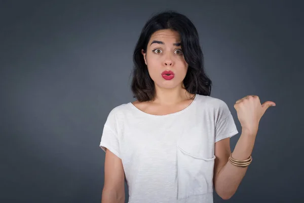 Horizontal shot of pretty young joyful shocked  woman  points with thumb away, indicates something on blank wall isolated over gray background. Check this out. Ethnicity concept.