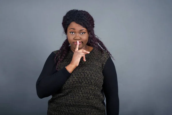 Dont tell my secret or not speak too loud, please! Emotional surprised student makes hush gesture, asks be quiet, has scared expression as afraids of revealing secret, poses against gray studio wall.