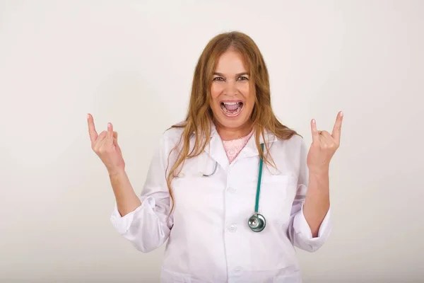 Born to rock this world. Joyful doctor woman in medical uniform screaming out loud and showing with raised arms horns or rock gesture, expressing excitement of being on concert of band.