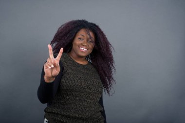 Gri duvara yaslanmış sıradan giyinmiş Afro-Amerikan kadın iki numaralı parmaklarıyla gülümserken kendinden emin ve mutlu..