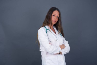 Kollarını kavuşturup tıbbi üniforma giymiş, gri duvara yaslanmış neşeli, beyaz bir doktor kadın resmi. Kameraya bakıp gülümsüyor. Kendine güvenen başarılı bir kadın..