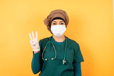 Yeşil üniformalı, yakışıklı, Müslüman doktor kadın duvarın karşısında duruyor ve üç numaralı parmaklarıyla gülümsüyor ve gülümsüyor..