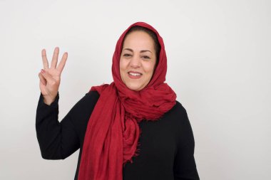 Olgun bir kadın gri duvarın önünde duruyor. Üç numaralı parmaklarıyla onu gösteriyor ve gülümsüyor. Kendinden emin ve mutlu. Stüdyoda poz veren başörtüsü takan orta yaşlı bir Arap kadın.