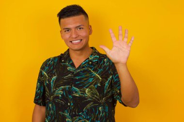 Genç İspanyol adam duvarın önünde duruyor ve beş numarayı gösterip işaret ederken kendinden emin ve mutlu bir şekilde gülümsüyor..