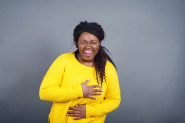 Sıradan giyinmiş neşeli Afro-Amerikalı kadın ellerini çapraz tutuyor, kapalı mekanda güzel şakalara gülüyor. Uzun saçlı mutlu genç kadın gri duvara poz veriyor..