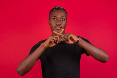 Genç, yakışıklı, Afro-Amerikan bir adam izole edilmiş arka planda günlük giysiler giyip duvarın önünde dikiliyor. Reddedilme ifadesi negatif işaret yaparak parmaklarını çapraz yapıyor mu?.