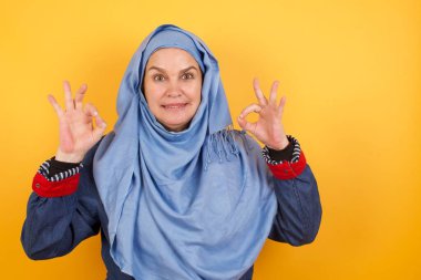 Orta yaşlı tesettürlü çekici müslüman kadın her iki elini de işaret olarak kullanıp neşeli bir ifade takınıyor.. 