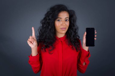 Kıvırcık saçlı, kırmızı tişörtlü, akıllı telefon gösteren ve gri stüdyoda parmağını gösteren genç, güzel esmer kadın. 
