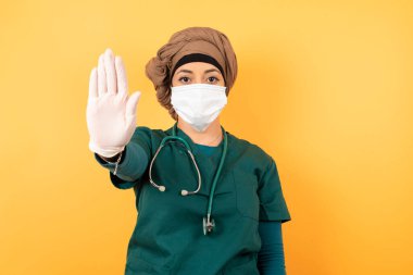 Yeşil üniformalı genç, güzel, Müslüman doktor kadın avucunun içi gibi jest yapıyor. Arka planda izole edilmiş yüz üzerinde negatif ve ciddi jest içeren uyarı ifadesi.