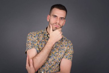 Düşünceli gülümseyen bir adamın portresi elini çenesinin altında tutar, doğrudan kameraya bakar, ilgiyle karışık bir şey dinler, sıradan giyinir, gri duvara karşı poz verir..