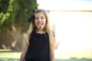 Gülümseyen ve dil gösteren mutlu ve komik yüzlü güzel beyaz kız. Günlük giysiler giyip parkta dikiliyor. 