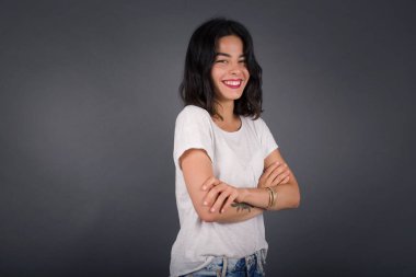 Kolları çapraz beyaz kazak giymiş, gri duvara yaslanmış neşeli beyaz kadın görüntüsü. Kameraya bakıp gülümsüyor. Kendine güvenen başarılı bir kadın..