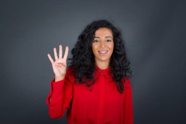 Kıvırcık saçlı, gri tişörtlü genç esmer kadın gri duvara yaslanmış ve parmaklarını dört numarayı gösterirken kendinden emin ve mutlu bir şekilde gülümsüyor..