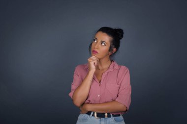 Düşünceli gülümseyen kızın portresi elini çenesinin altında tutuyor, yan bakıyor, ilgi çekici bir şey düşünüyor ya da merak ediyor, sıradan giyiniyor, gri stüdyoya karşı poz veriyor..