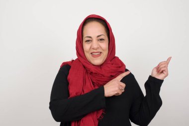 Reklam konsepti. Gri arka planda kot gömlekli, ışıl ışıl gülümseyen kadın işaret parmağıyla işaret ediyor ve boş fotokopi alanına bakıyor. Stüdyoda poz veren başörtüsü takan orta yaşlı bir Arap kadın.