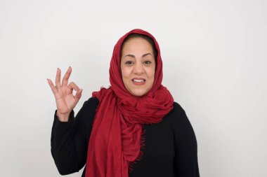 Olgun Müslüman bir kadının, olumlu bir ifadeyle, olumlu bir ifadeyle, olumlu bir ifadeyle, el işaretini göstermesine sevindim. Beyaz duvara yaslanarak.