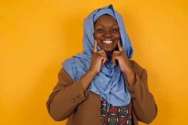 Güçlü, sağlıklı, beyaz dişlere sahip olma kavramı. Arka planda izole edilmiş, kusursuz beyaz dişlerini gösteren ışıl ışıl gülümseyen mutlu Afro-Amerikalı Müslüman kadının portresini yakından çek..