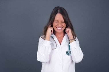 Bu sinir bozucu sesi çıkarmayı kes! Stresli genç bir doktor kadın, parmaklarıyla kulaklarını tıkamış, üst katındaki komşularından gelen gürültüden rahatsız olmuş..