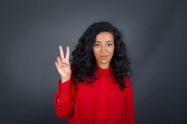 Kıvırcık saçlı, gri tişörtlü genç esmer kadın gri duvarın karşısında duruyor ve iki numaralı parmaklarıyla işaret ediyor kendinden emin ve mutlu bir şekilde gülümsüyor..