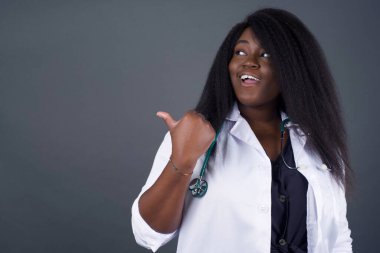 Omg konsepti. Şaşırmış Afrika kökenli Amerikalı kadın doktor, gözlerini ve ağzını iyice açıyor, baş parmağıyla işaret ediyor, gri arka planda garip bir şey gösteriyor. Reklam kavramı.