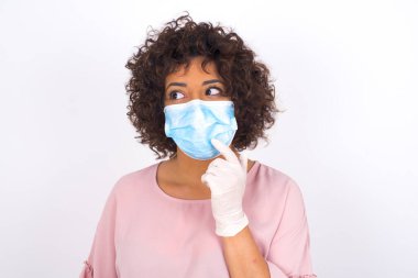 Yüz maskesi takan ve günlük kıyafet giyen bir kadın beyaz stüdyo arka planında izole edilmiş olduğunu düşünüyor.