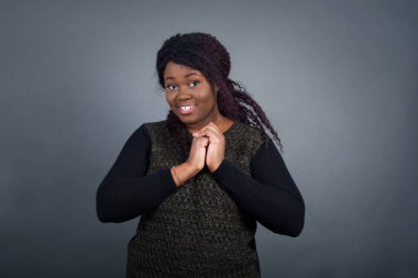 Hoş yüzlü, rüya gibi, Afro-Amerikalı bir kadın. Elleri çapraz, hoş bir şey için heyecanlı, gri arka planda poz veriyor..