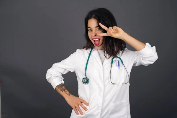 Leisure lifestyle people person celebrate flirt coquettish concept. Beautiful doctor woman wearing medical uniform shows v-sign near eyes standing against gray wall.