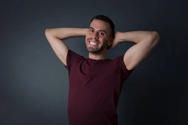 Confidence and coquettish concept. Portrait of charming young man, smiling broadly with self-assured expression while holding hands over her head. Standing against gray background.