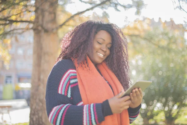 Güz parkında tablet kullanan uzun saçlı güzel Afro-Amerikan kadın renkli kazak giyiyor.  