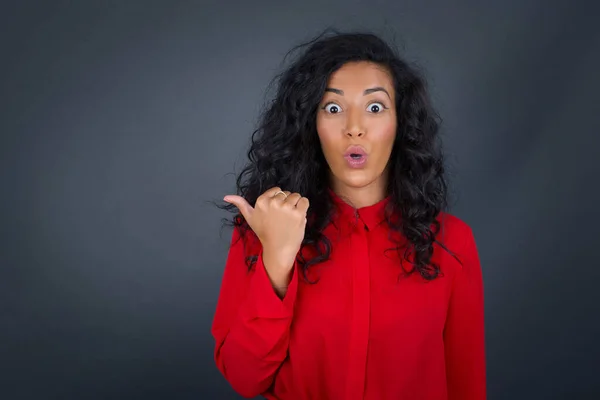 Horizontal shot of pretty young joyful shocked brunette woman with curly hair wearing red shirt points with thumb away, indicates something on blank wall isolated over gray background.