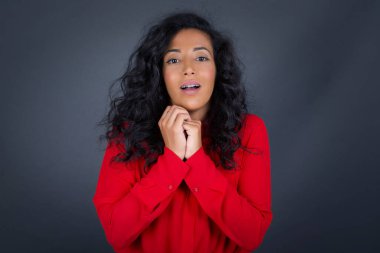 Kıvırcık saçlı, kırmızı tişörtlü, rüya gibi genç esmer kadın ellerini yüzünün yanında gezdiriyor, hoş bir şey için heyecanlı, gri arka planda poz veriyor..