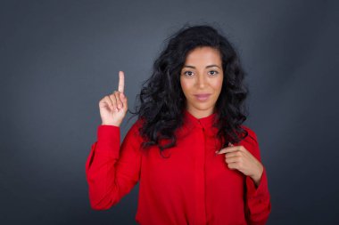 Kıvırcık saçlı, kırmızı tişörtlü, ağzı açık neşeli esmer kadın gri stüdyoda bir şeyler gösteriyor. Bir eli göğsünde ve diğer eliyle işaret ediyor.. 