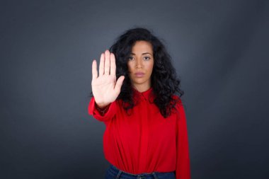 Kıvırcık saçlı, kırmızı gömlekli genç, güzel esmer kadın avuç içiyle jest yapıyor. Gri arkaplan üzerinde izole edilmiş yüz üzerinde negatif ve ciddi jest içeren uyarı ifadesi.