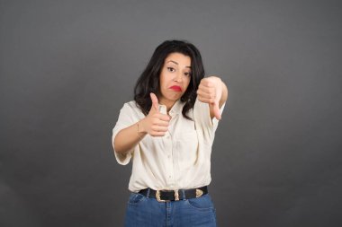 Güzel esmer kadın, baş parmağı yukarıda, baş parmağı aşağıda. Gri arkaplanda duruyor. 