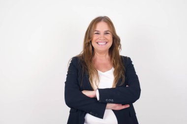 Orta yaşlı, beyaz, mutlu, kollarını kavuşturmuş gülümseyen bir kadın gri duvara yaslanmış kameraya kendinden emin bir şekilde bakıyor. Pozitif kişi.